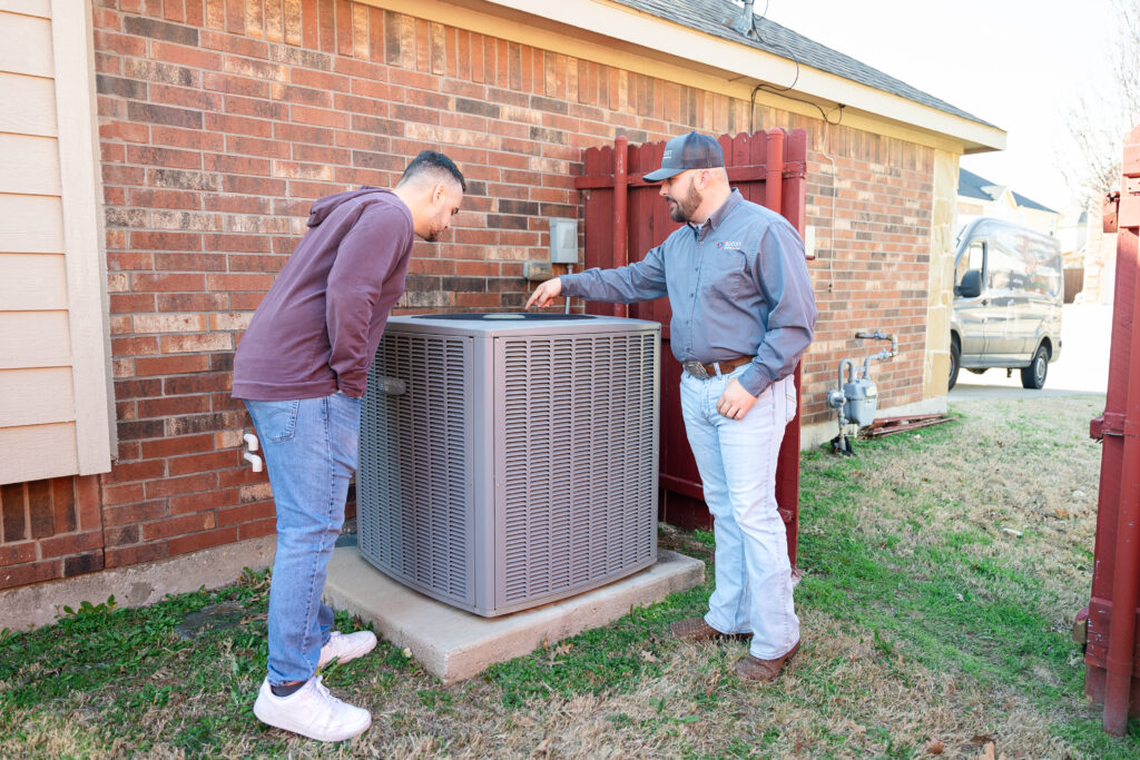 Boost Air technician inspecting Ruud outdoor AC condenser with homeowner by brick house
